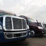 Grilles and headlights of modern semi trucks like the eyes and mouth of huge monsters, bristling gloss chrome lined up on a huge truck stop site against a gray stormy cloudy sky.